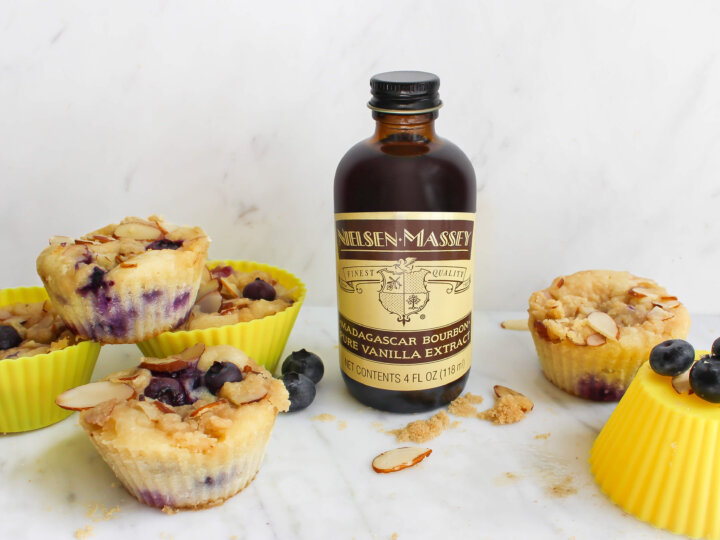Blueberry muffins in yellow muffin papers on a counter next to a bottle of Nielsen-Massey Madagascar Bourbon Pure Vanilla Extract